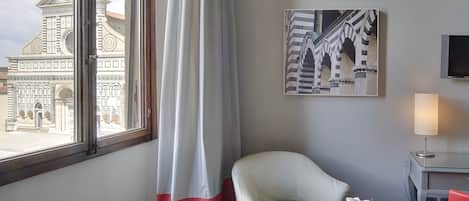 Habitación de hotel con dos sillones tapizados, una mesita redonda con un frutero, una lámpara y un cuadro de un edificio en la pared.