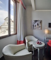 A hotel room with two upholstered chairs, a small round table with a bowl of fruit, a lamp, and a painting of a building on the wall.