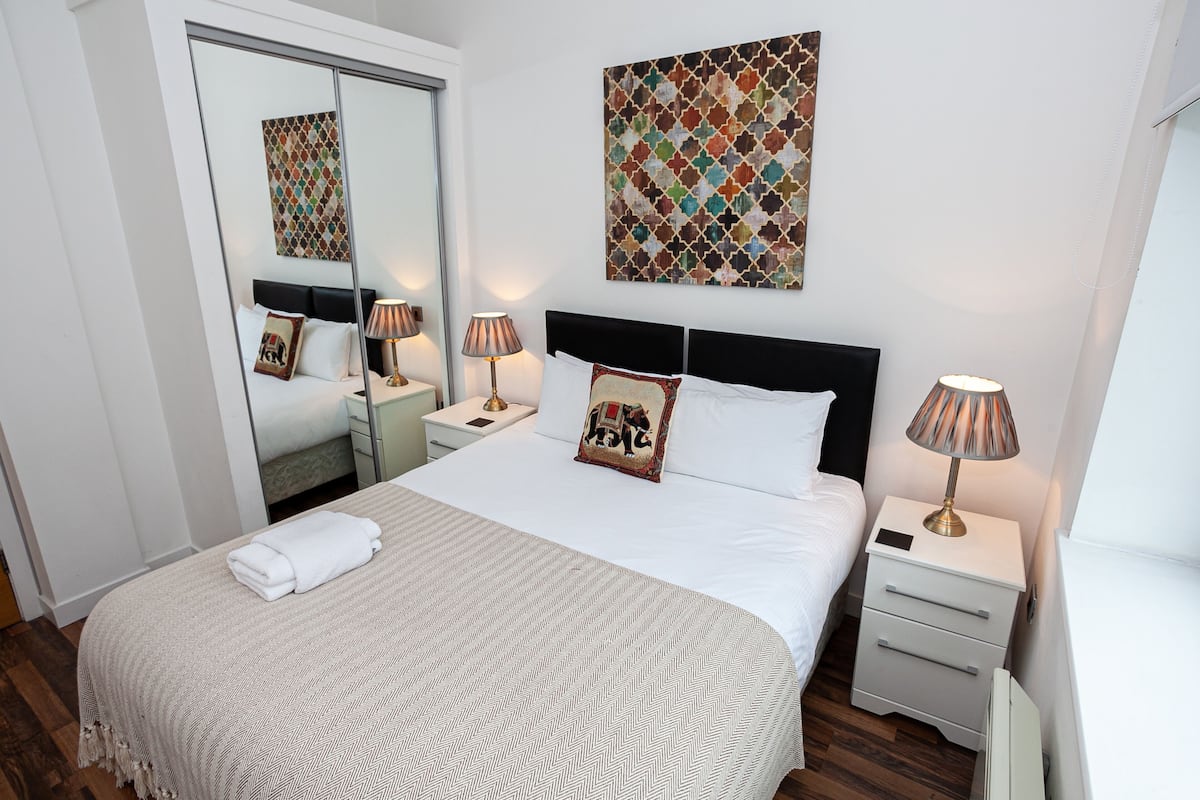 Een net opgemaakt bed met een witte deken en een gevouwen witte handdoek, twee nachtlampjes, een zwart hoofdeinde, een spiegel en een kunstwerk met een geometrisch patroon aan de muur.