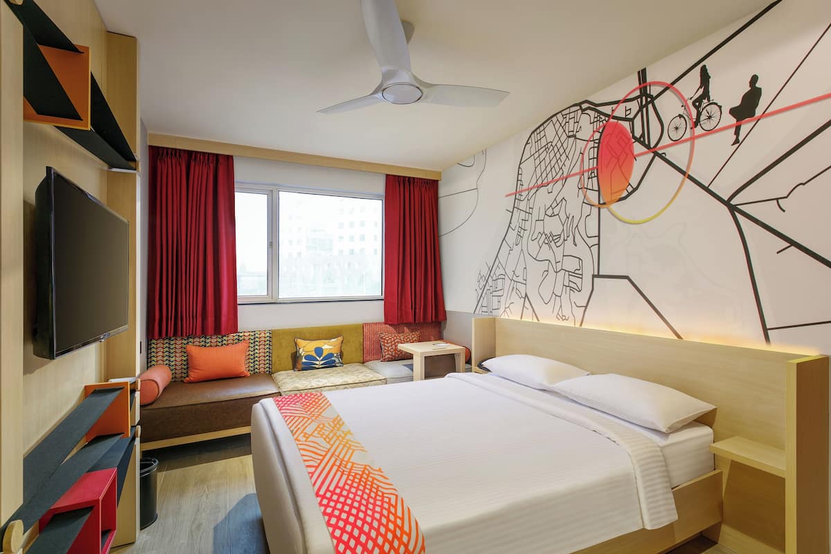 A hotel room with a bed, a TV, a desk, a chair, a sofa, and a window with curtains.