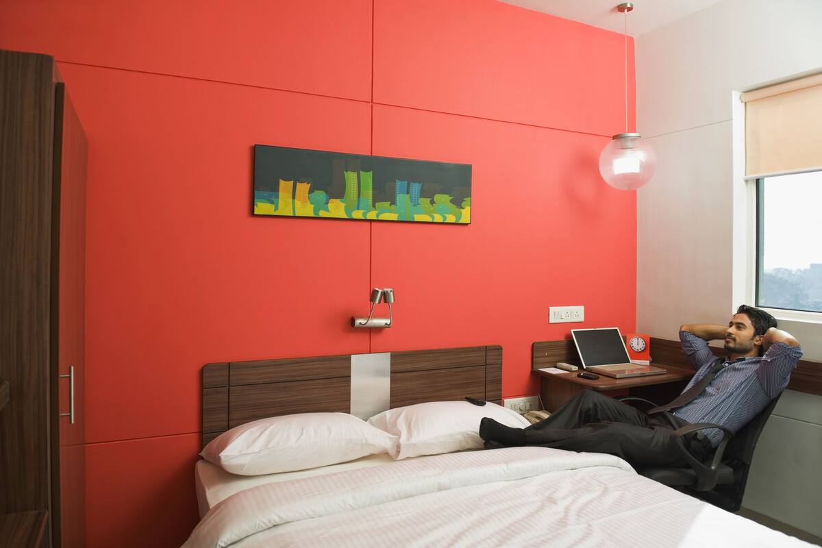 A hotel room with a red wall, a bed with white bedding, a desk with a laptop, and a cityscape painting.
