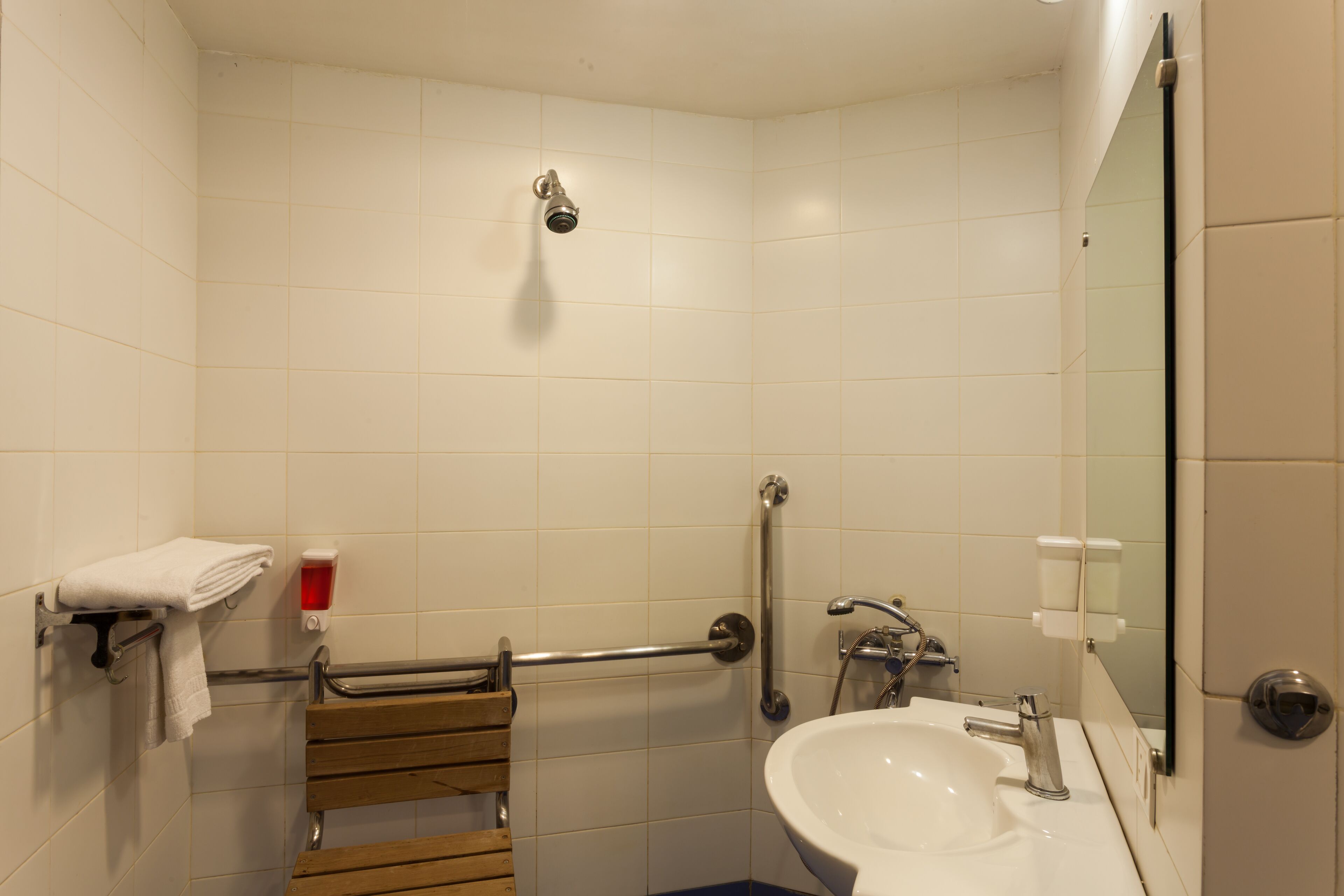 Un baño con azulejos blancos, un lavamanos, un espejo, una ducha, un perchero para toallas y una barra de apoyo.