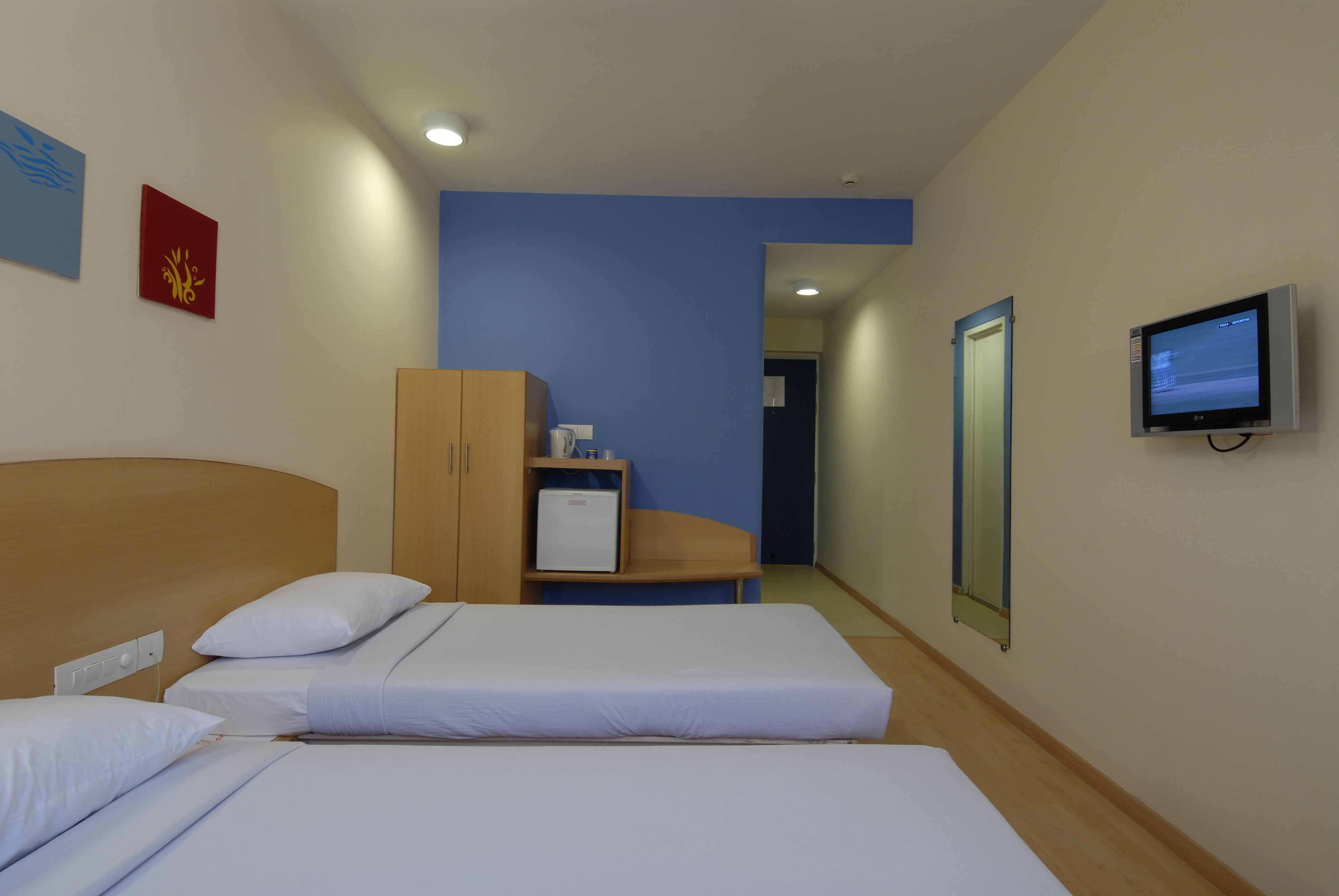 Une chambre d’hôtel avec deux lits, une armoire en bois, un petit réfrigérateur, une télévision et une porte donnant sur une autre chambre.