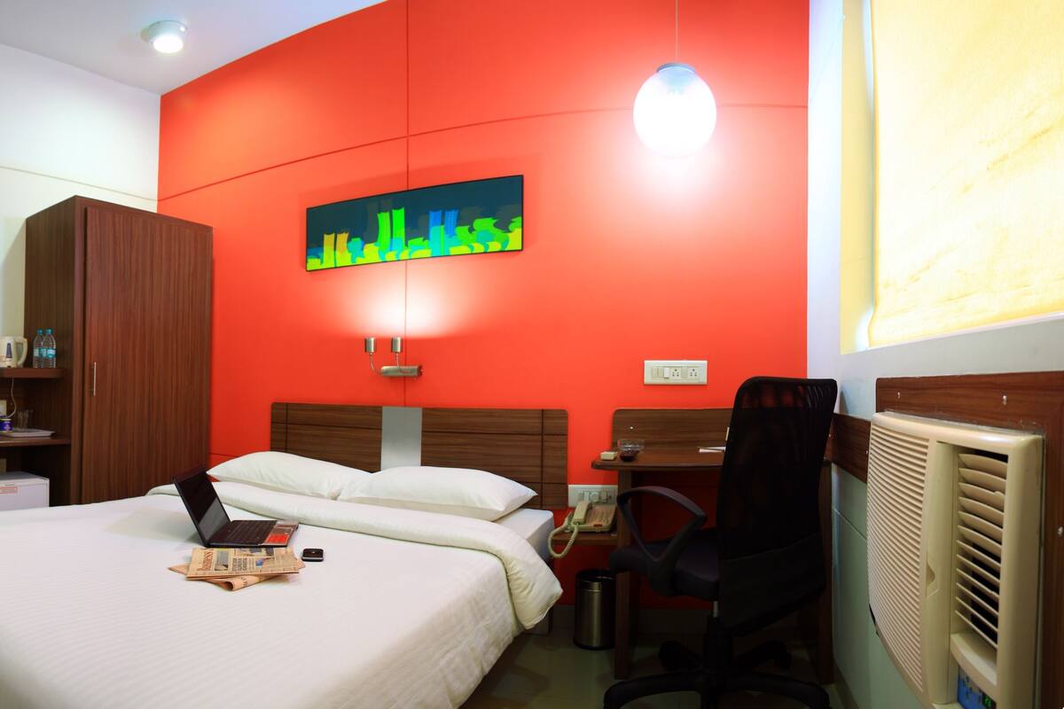 A hotel room with a bed, a desk with a laptop, a chair, a radiator, and a TV on the wall.