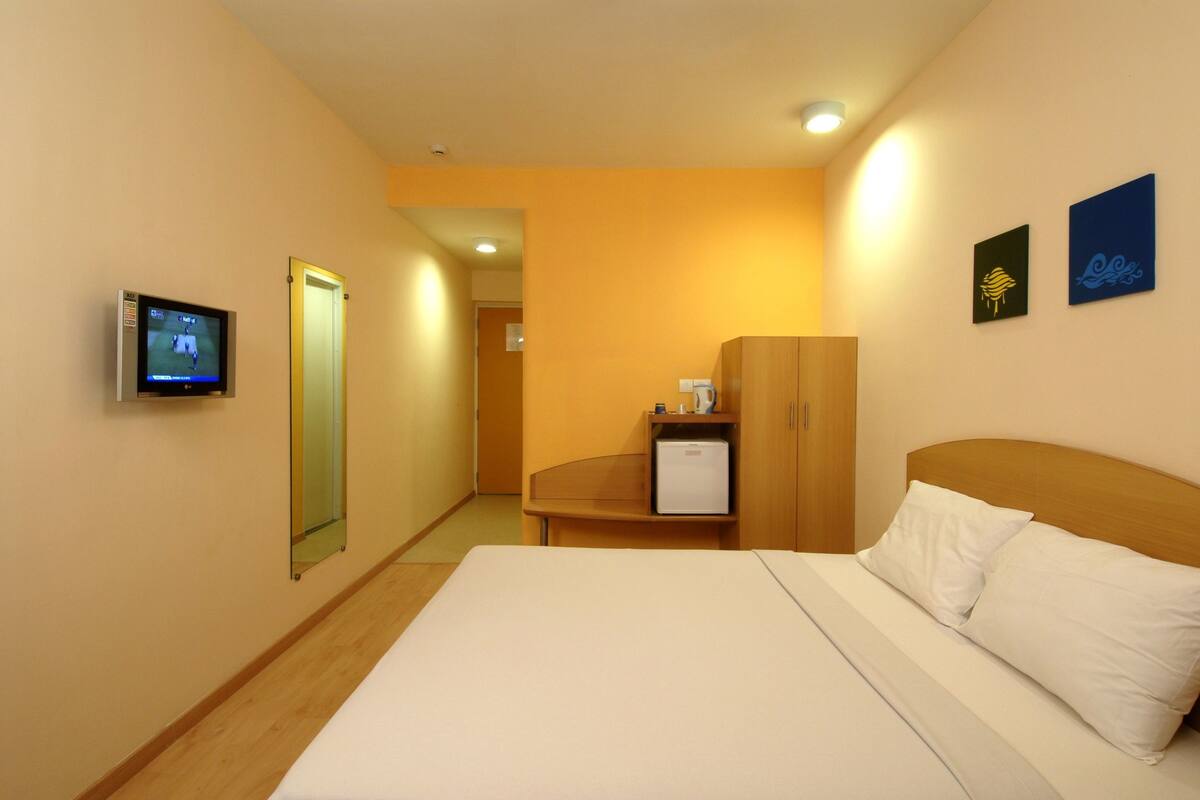 A hotel room with a bed, a television, a wardrobe, and a small desk with a refrigerator.