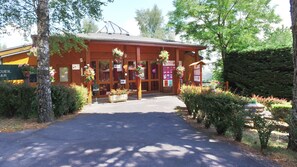 A wooden building with a covered entrance, surrounded by greenery and trees. - VVF Aubrac, Chaudes-Aigues (Chaudes-Aigues)
