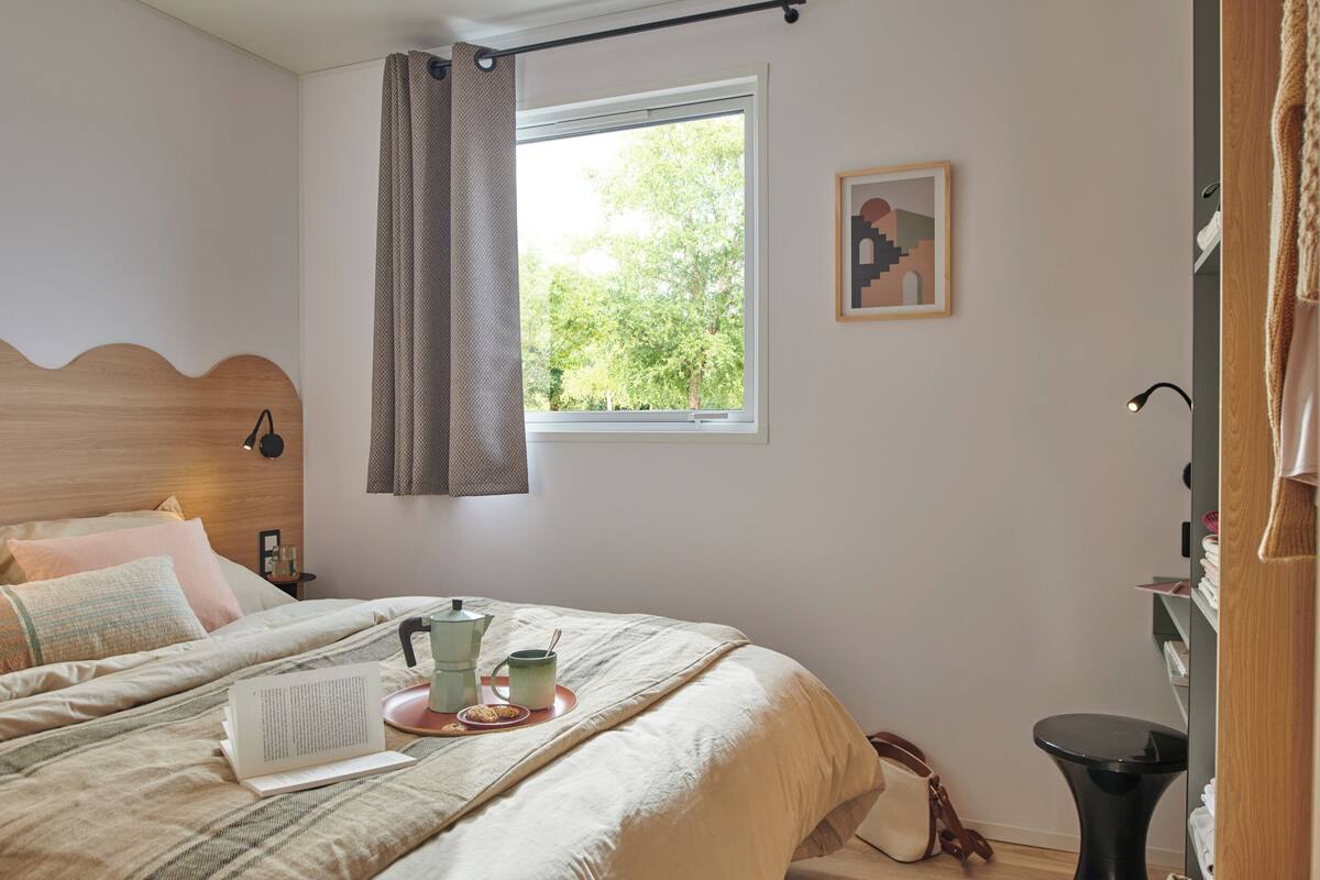 Ein Schlafzimmer mit Bett, einem Fenster mit Vorhängen, einem Nachttisch, einem gerahmten Bild und einem Beistelltisch.