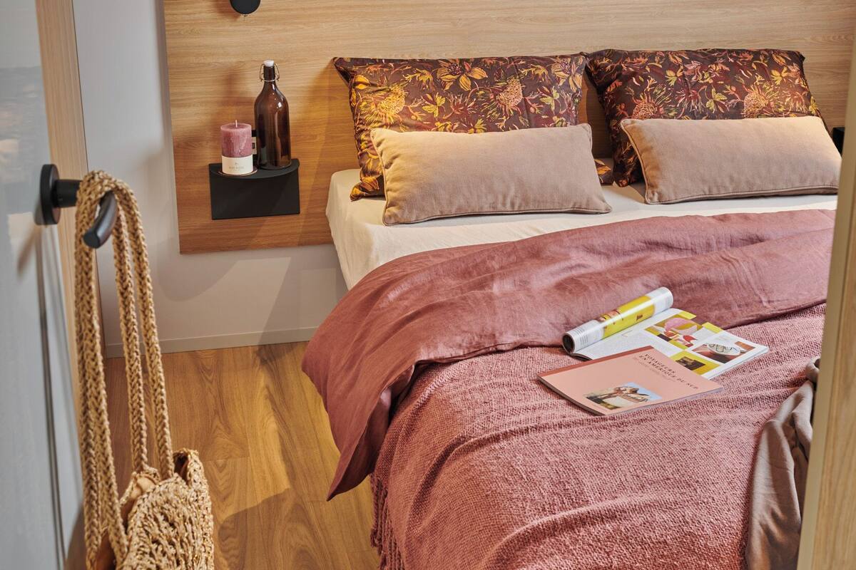 Ein ordentlich bezogenes Bett mit einer rosa Decke, einem hölzernen Kopfteil und einem Nachttisch mit Kerze und Flasche.