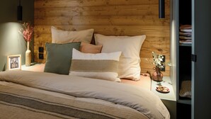 A bedroom with a wooden headboard, a bed with multiple pillows, a bedside table with a vase and a picture frame, and a hanging light fixture. - VVF Aubrac, Chaudes-Aigues (Chaudes-Aigues)