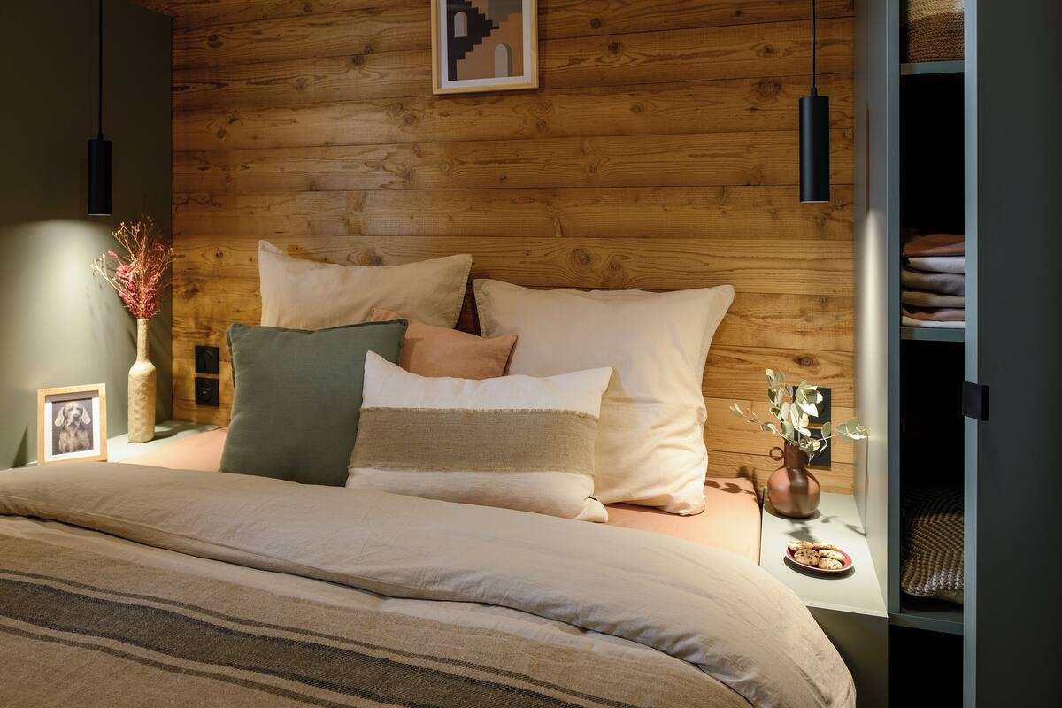Ein Schlafzimmer mit Holz-Kopfteil, einem Bett mit vielen Kissen, einem Nachttisch mit Vase und Bilderrahmen sowie einer Pendelleuchte.