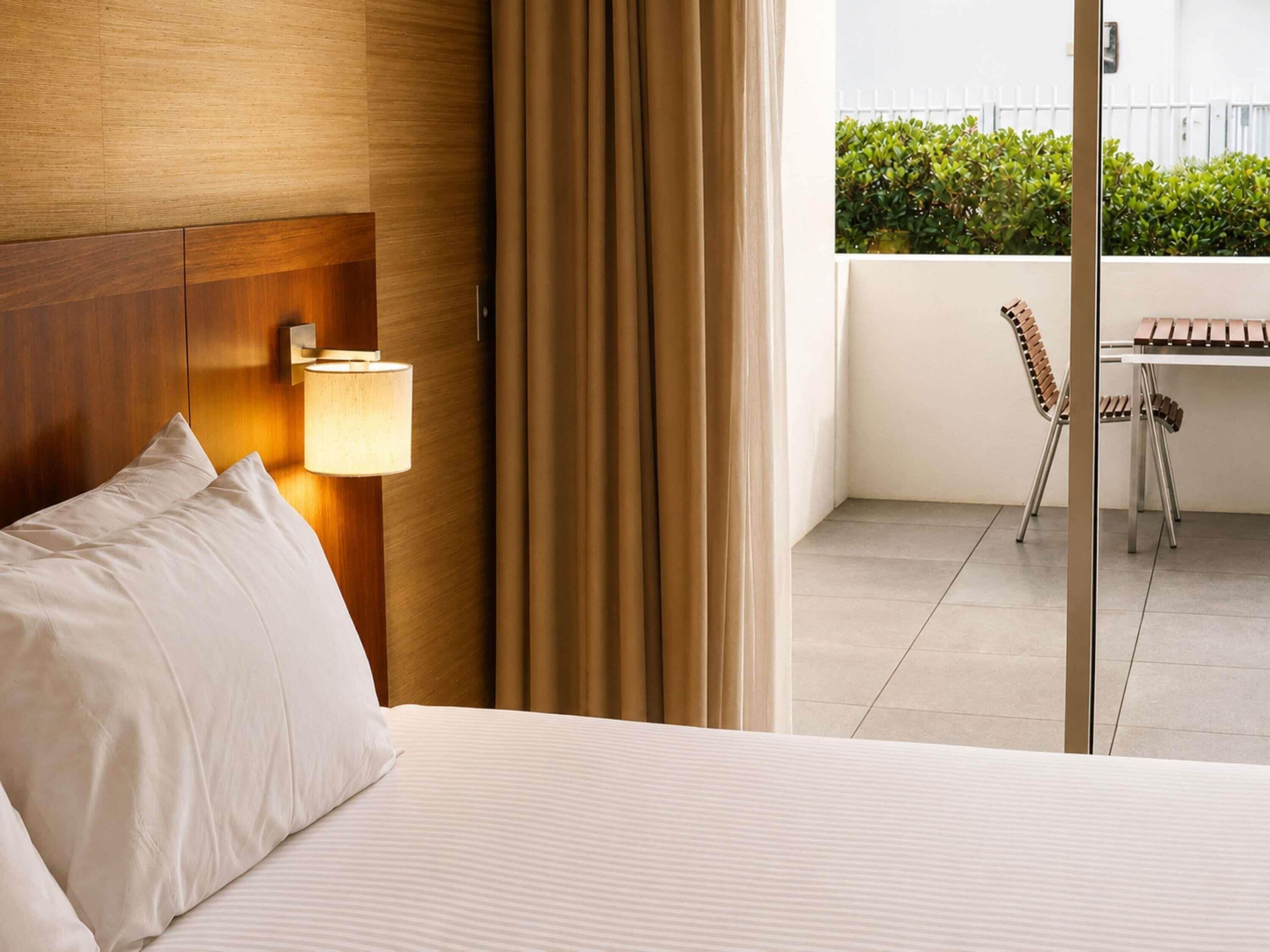 Ein Hotelzimmer mit Bett, Lampe, Blick auf einen Balkon mit Stuhl und Tisch sowie einer weißen Wand.