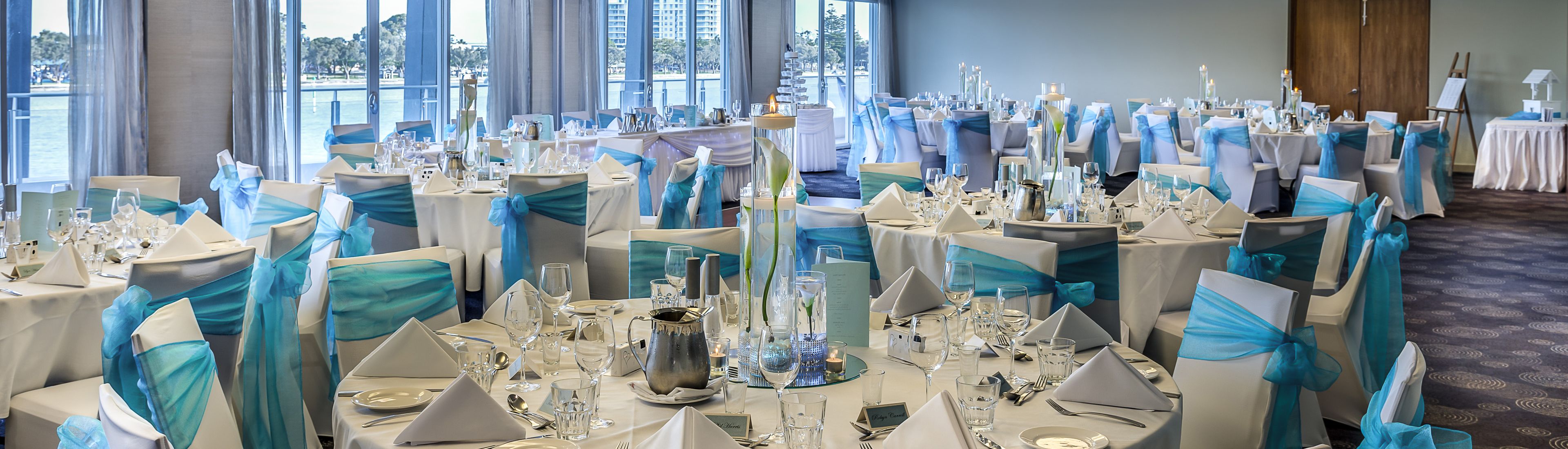 Un salón de banquetes con mesas redondas dispuestas para un evento formal, con cintas color turquesa y manteles blancos.