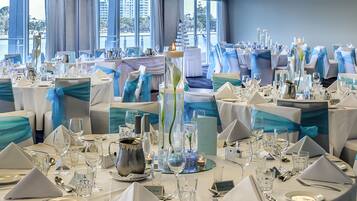 Un salón de banquetes con mesas redondas dispuestas para un evento formal, con cintas color turquesa y manteles blancos.