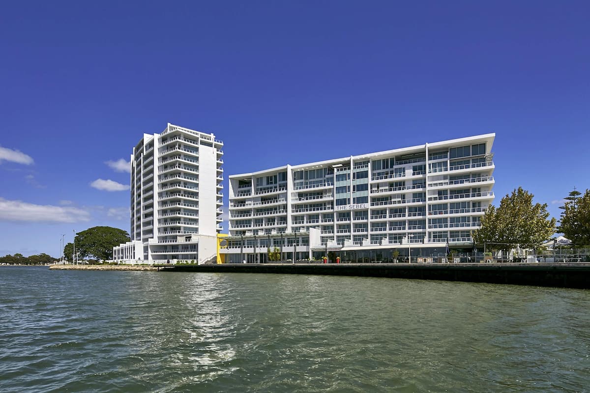 A modern hotel with multiple floors, a waterfront view, and a prominent sign reading "THE SEBEL".