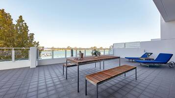 A rooftop terrace with a view of the sea, a wooden table, two blue lounge chairs, and a metal bench.