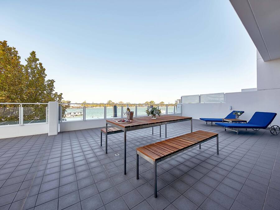 A rooftop terrace with a view of the sea, a wooden table, two blue lounge chairs, and a metal bench.