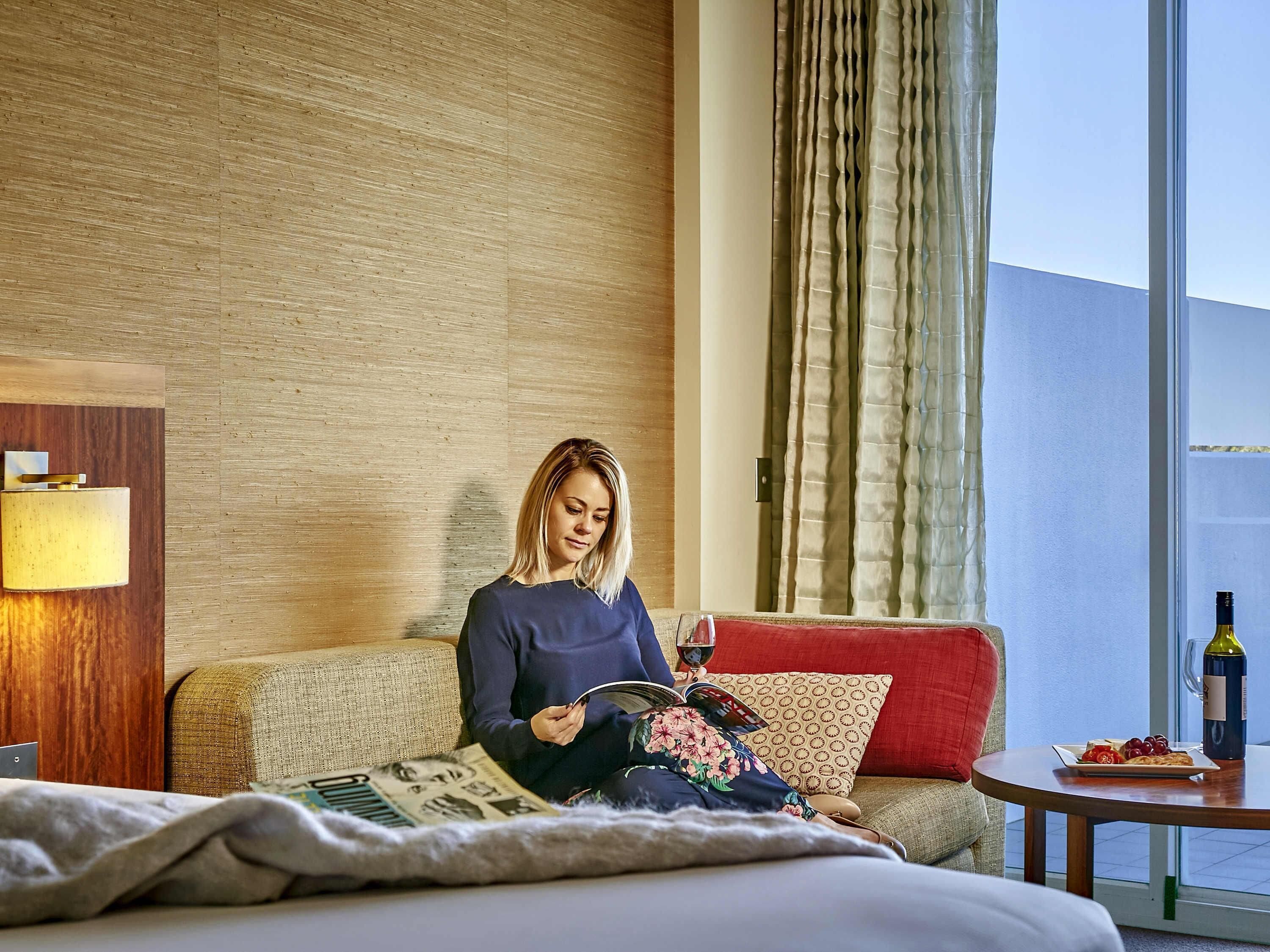 Una habitación de hotel con una cama, un sofá, una mesita con una botella y un plato de uvas, y una ventana con cortinas.