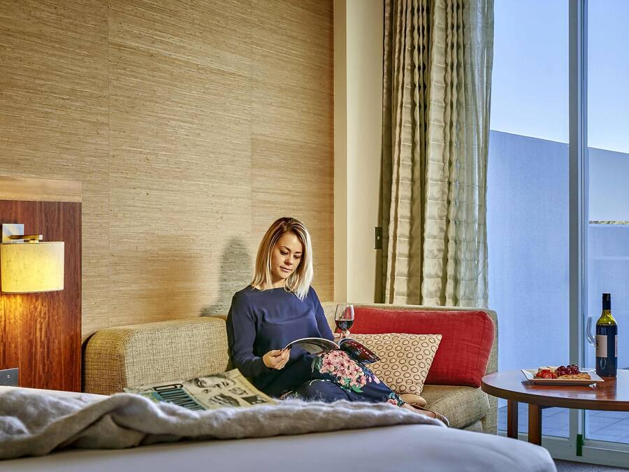 Una habitación de hotel con una cama, un sofá, una mesita con una botella y un plato de uvas, y una ventana con cortinas.
