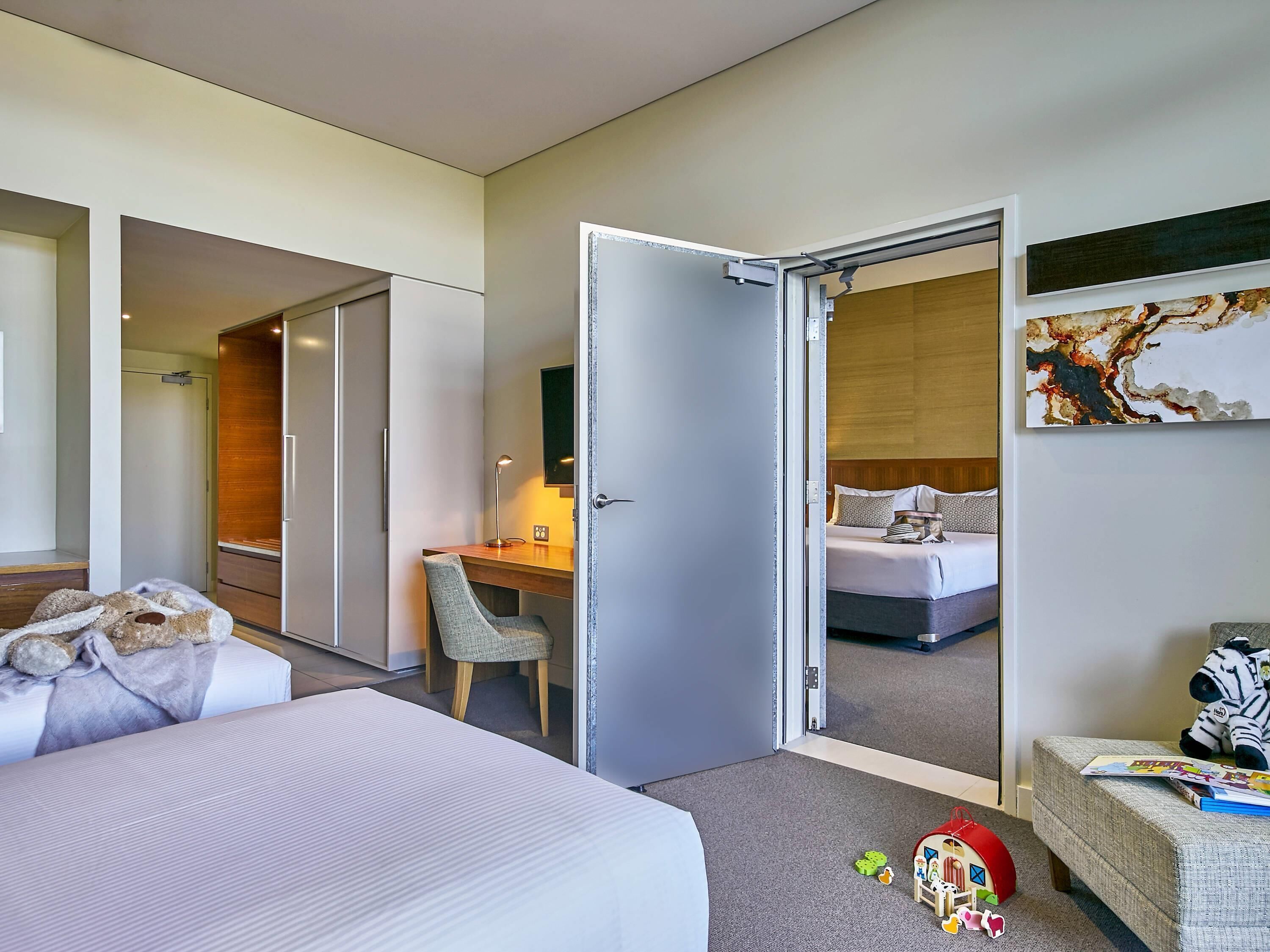 Habitación de hotel con cama, escritorio, silla, pequeña mesa con lámpara, puerta que da a otra habitación y un cuadro en la pared.