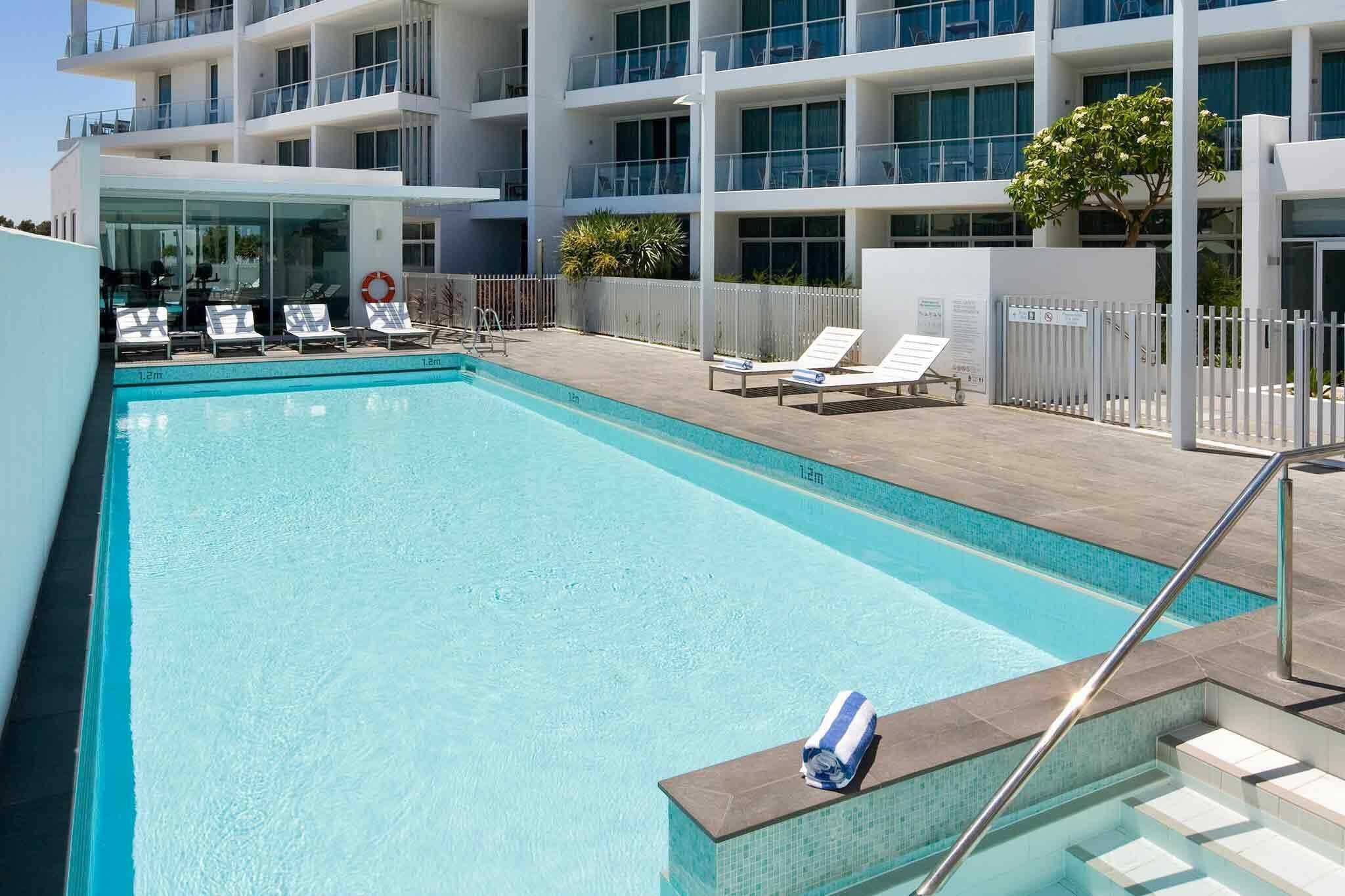 Una piscina con sillas de descanso, un flotador salvavidas y un edificio con varios balcones al fondo.