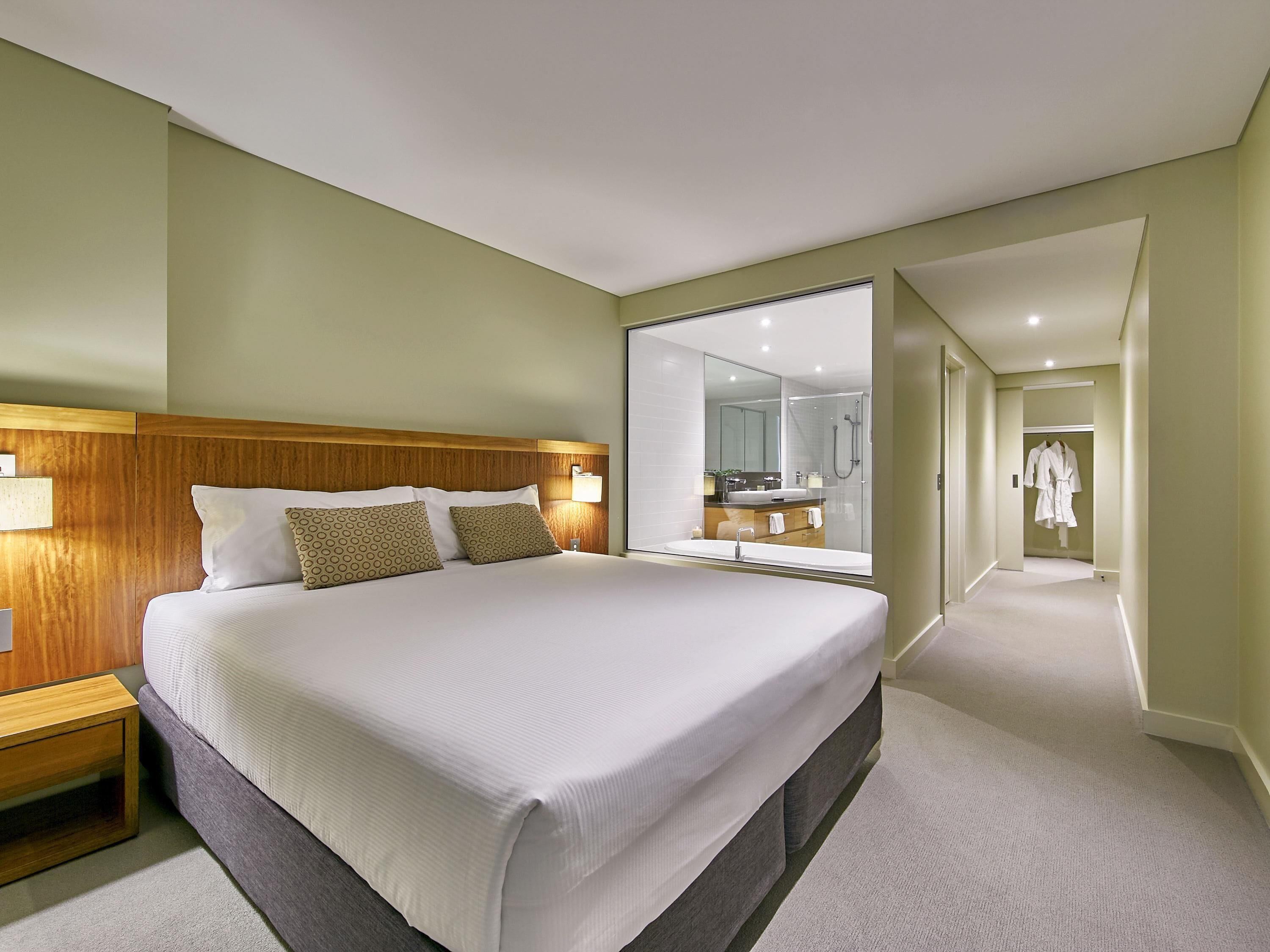 A hotel room with a large bed, a wooden headboard, a bathroom with a large mirror, and a walk-in closet with a robe hanging.