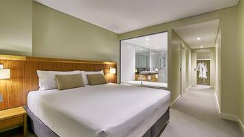 A hotel room with a large bed, a wooden headboard, a bathroom with a large mirror, and a walk-in closet with a robe hanging.