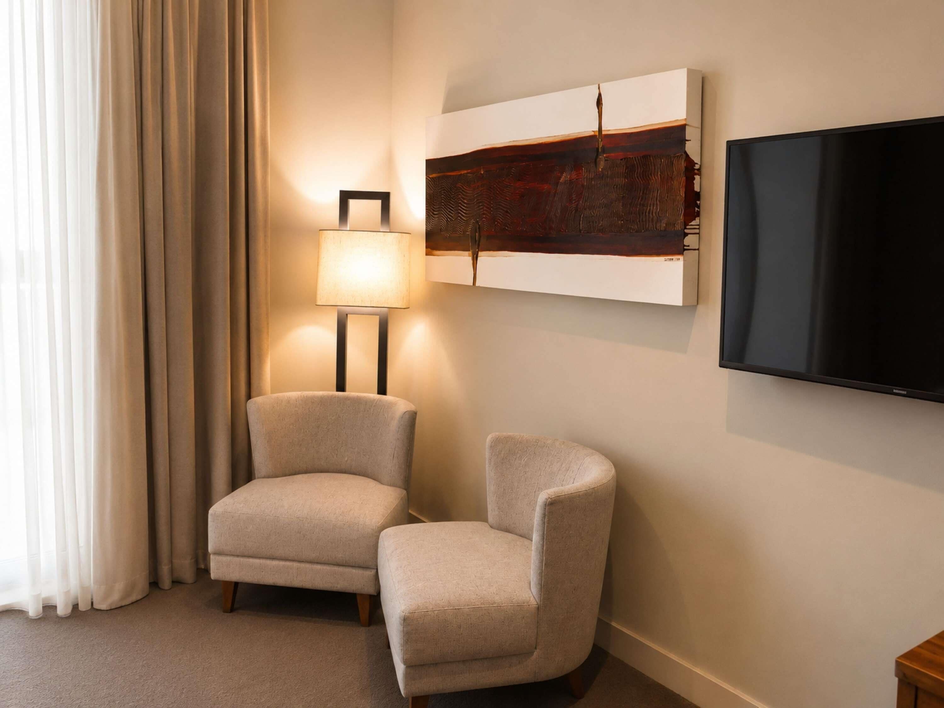 A hotel room with a flat-screen TV mounted on the wall, two armchairs, a floor lamp, and a large abstract painting.