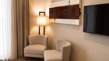 A hotel room with a flat-screen TV mounted on the wall, two armchairs, a floor lamp, and a large abstract painting.