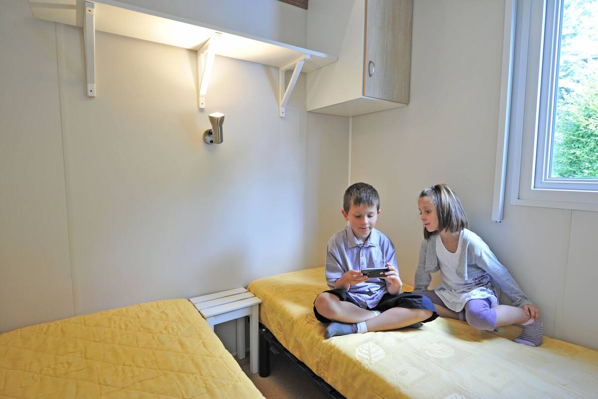 Ein Zimmer mit zwei Betten, eines mit gelber Bettwäsche und das andere mit weißer Bettwäsche. Rechts befindet sich ein Fenster, darüber eine Ablagefläche.