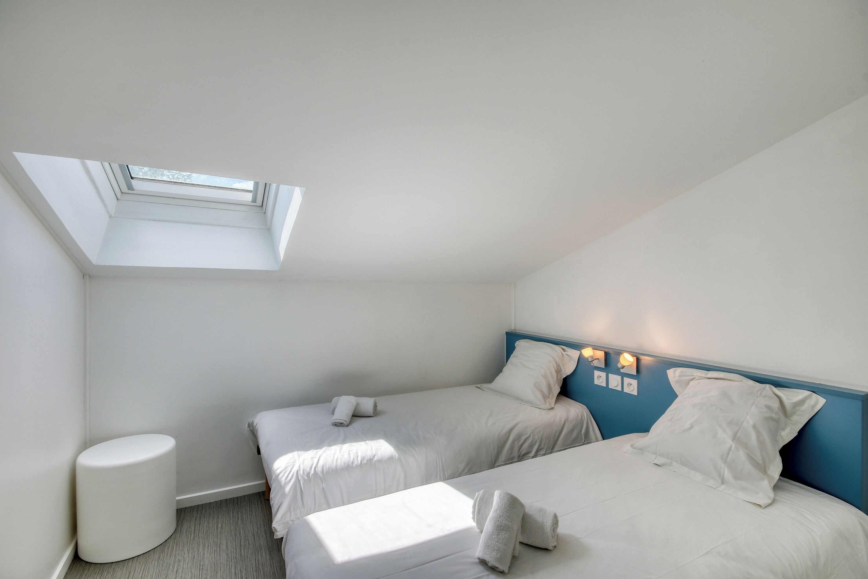 Habitación con dos camas, un tragaluz y un taburete blanco pequeño.