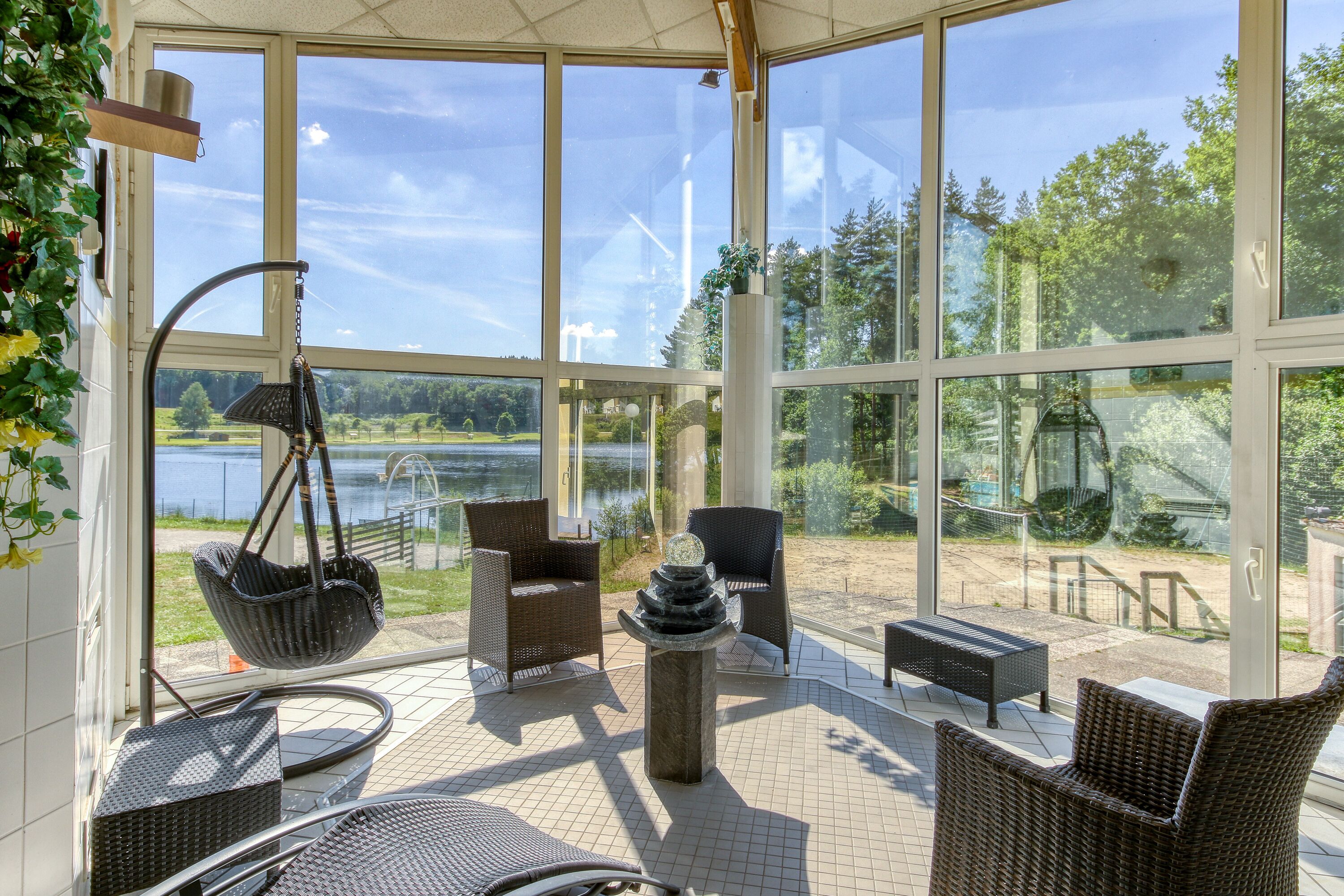 Une terrasse ensoleillée, meublée de mobilier en osier, avec un fauteuil suspendu et une vue sur un lac.