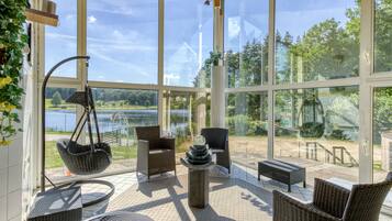 Une terrasse ensoleillée, meublée de mobilier en osier, avec un fauteuil suspendu et une vue sur un lac.