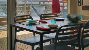 A dining area with a table set for two, a woman in a red dress standing by a glass door, and a view of a lake and trees. - VVF Corrèze Pays d'Eygurande (Eygurande)