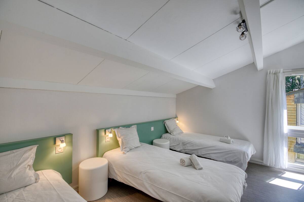Ein Zimmer mit zwei Betten, einem kleinen runden Tisch und einem Fenster mit Vorhängen.