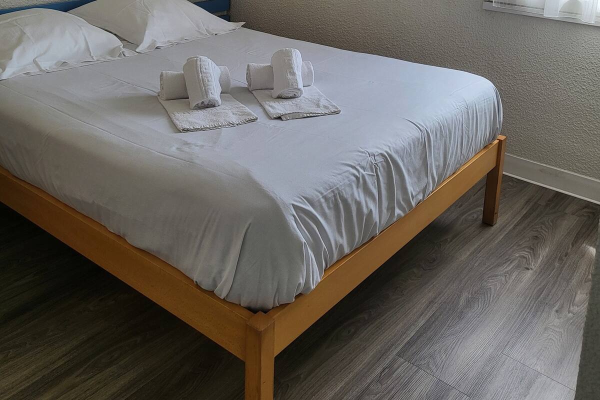 A neatly made bed with white linens and towels, a wooden headboard, a wall-mounted lamp, and a window with sheer curtains.