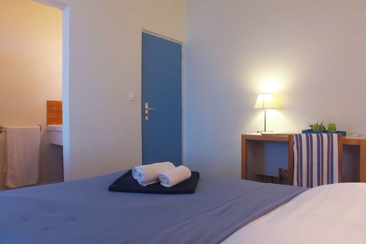 A hotel room with a bed, a desk with a lamp, and a door.