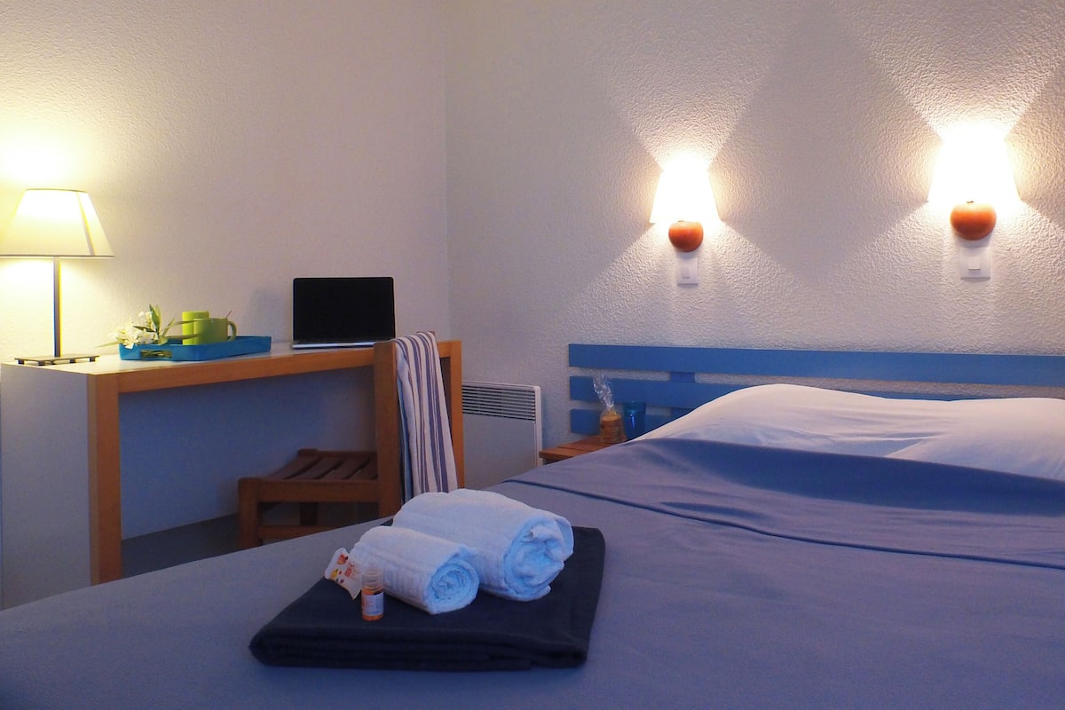 A hotel room with a bed, a desk with a laptop, a lamp, and two wall-mounted lights.