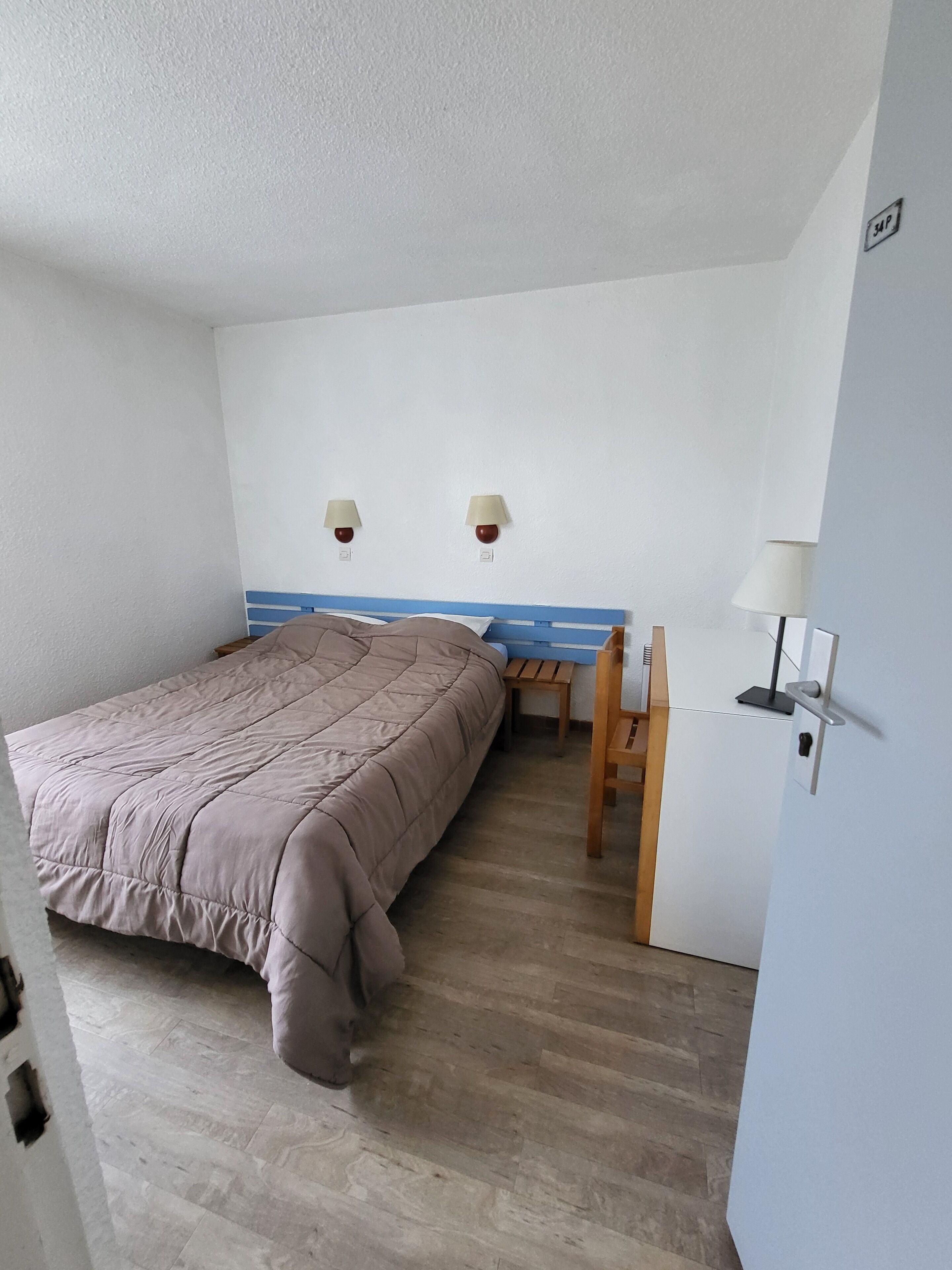 Une chambre à coucher avec un lit, un bureau et une chaise. Il y a deux lampes fixées au mur et une porte avec une pancarte.