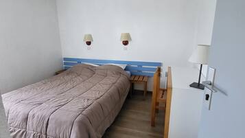 Une chambre à coucher avec un lit, un bureau et une chaise. Il y a deux lampes fixées au mur et une porte avec une pancarte.