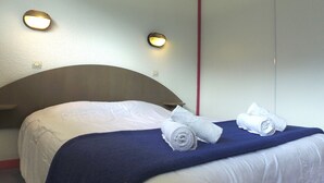 A neatly made bed with white linens and a blue blanket, two rolled towels, and two wall-mounted lights. - VVF Queyras, Ceillac-en-Queyras (Ceillac)