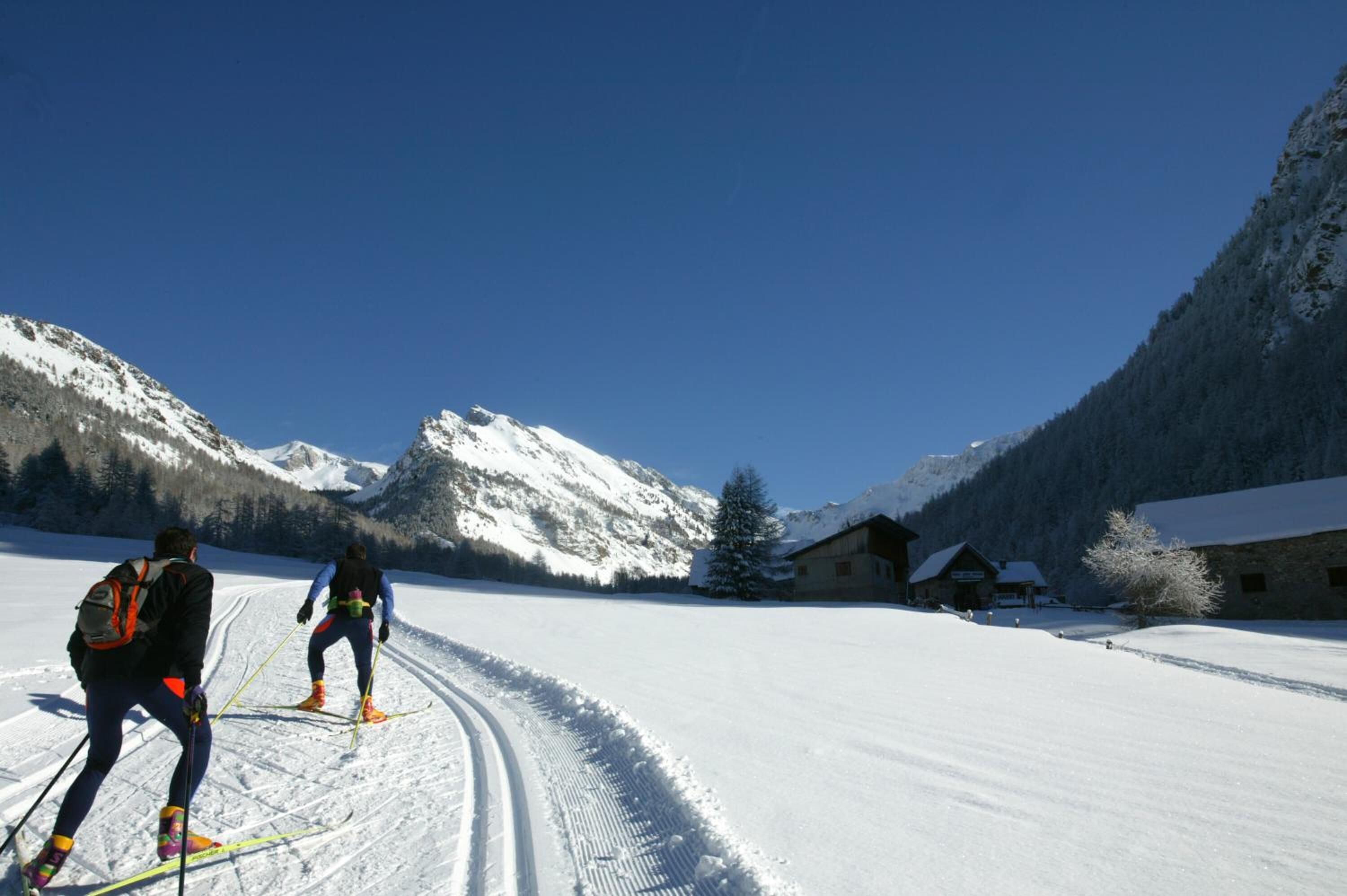Deux skieurs sur une piste enneigée, avec une montagne en arrière-plan et un bâtiment visible au loin.