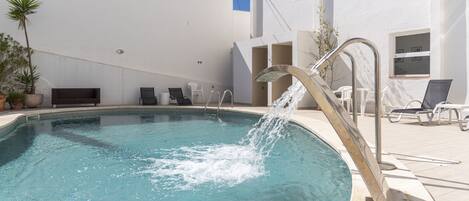 Una piscina con fuente, rodeada por un edificio blanco de techo plano, con mobiliario exterior y plantas en macetas.