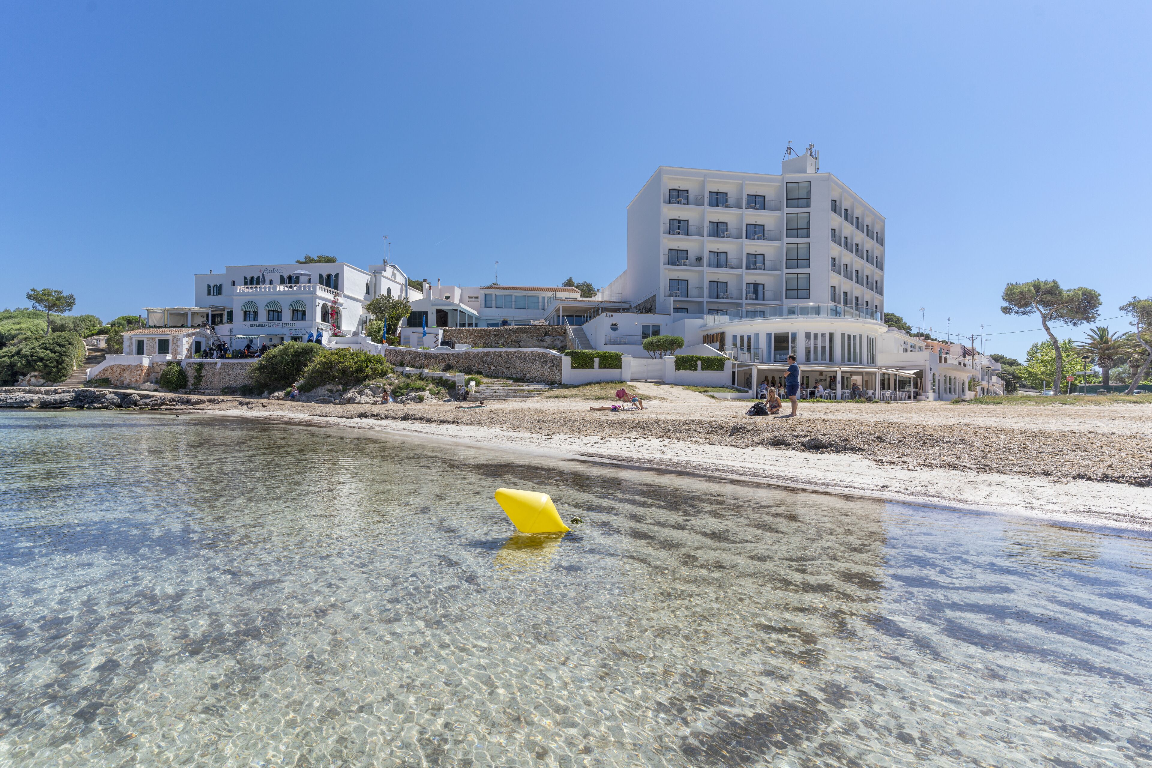 A coastal hotel with multiple buildings, clear waters, and a sandy beach.