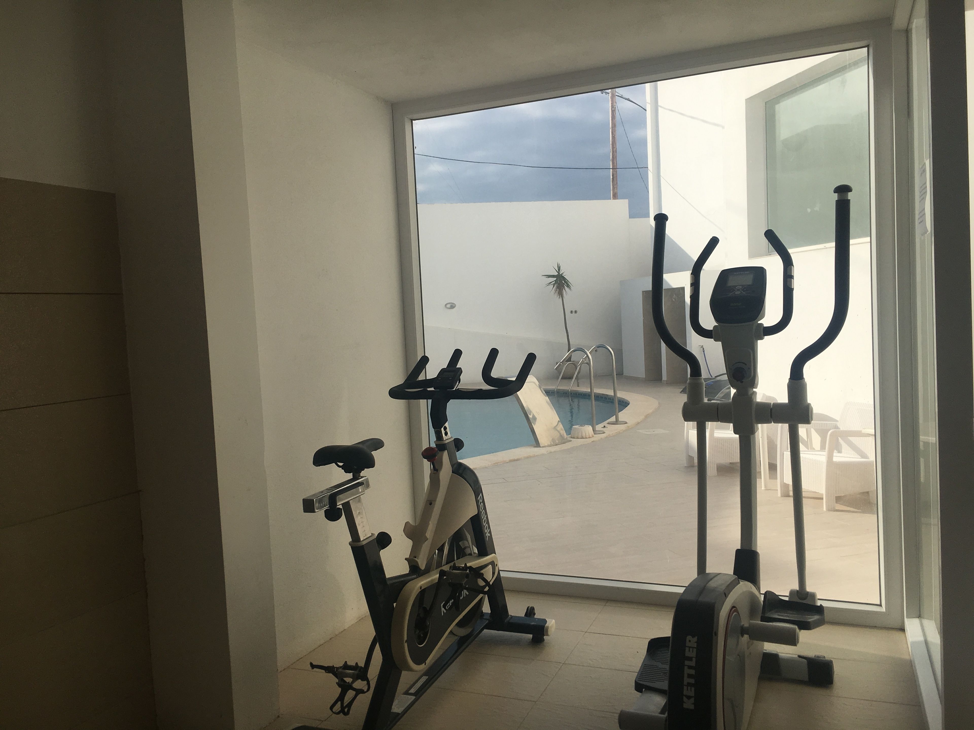 Ein Fitnessraum mit Ergometer, Crosstrainer und Laufband, ein kleiner Pool und Blick auf Himmel und Wolken.