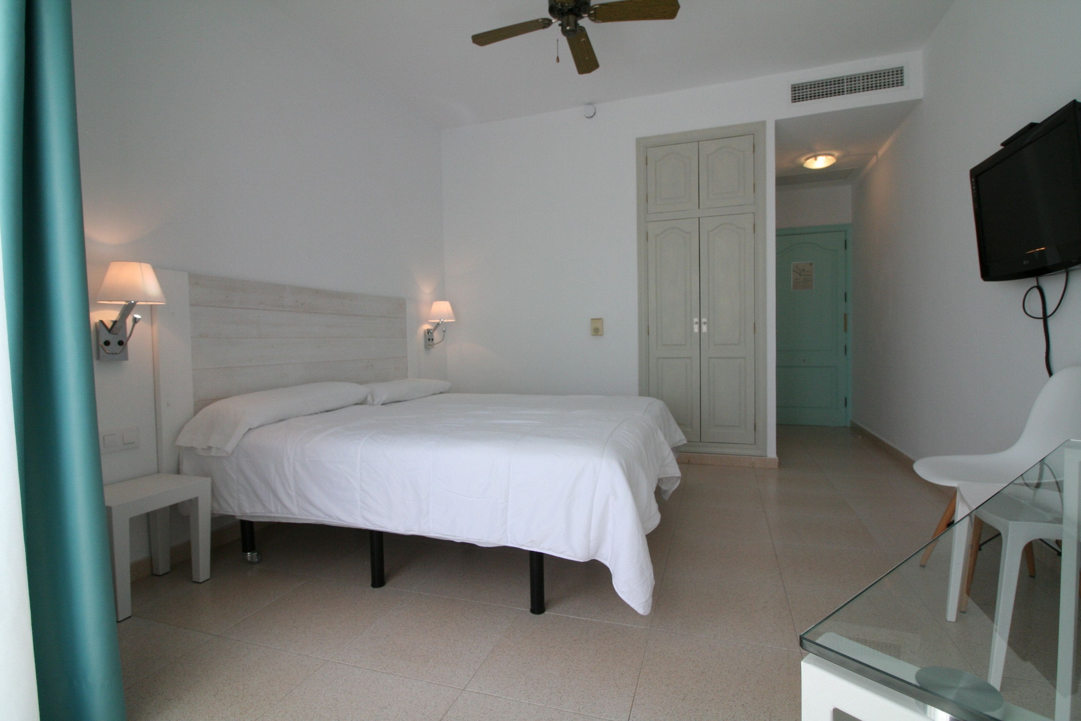 Une chambre à coucher comprenant un lit, des tables de chevet, un ventilateur de plafond, une télévision et une porte donnant sur une autre pièce.