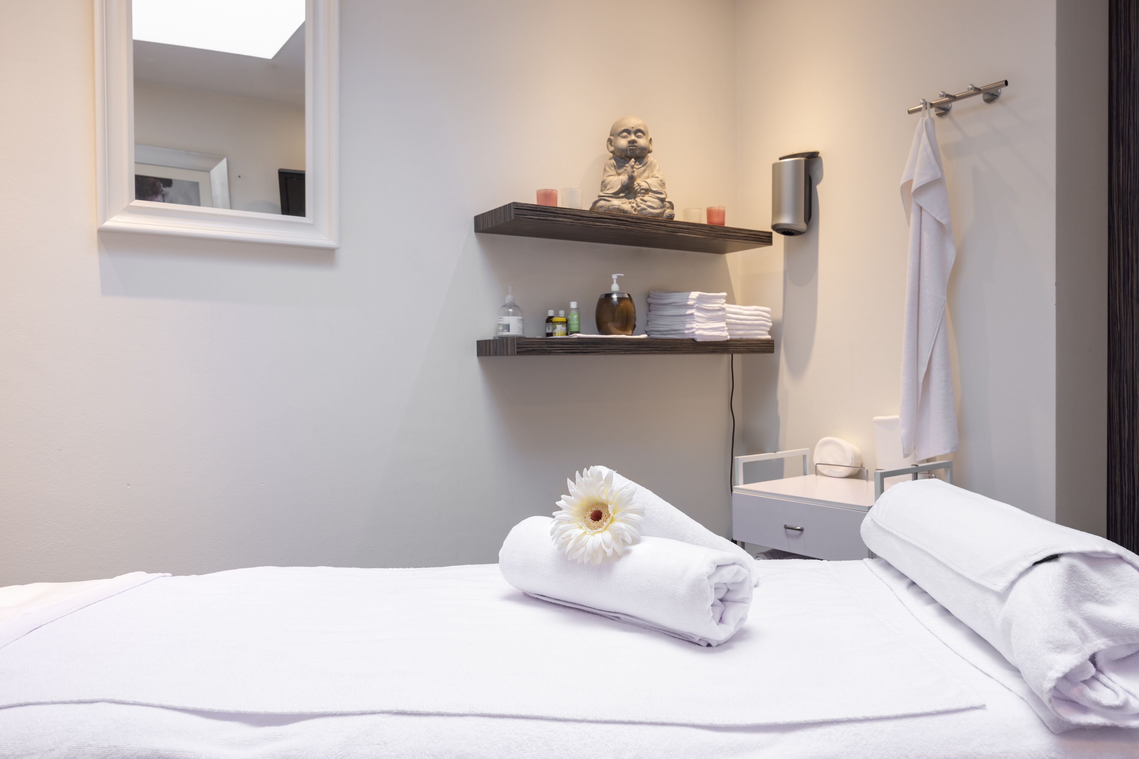 A hotel room with a bed, a nightstand, a mirror, a shelf with toiletries, and a towel with a flower.