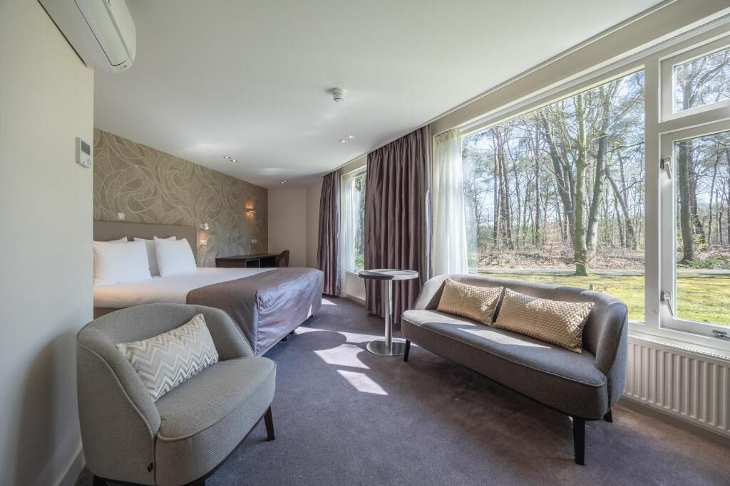 A hotel room with a bed, a sofa, a chair, and a small table. There is a window with curtains and a view of trees.