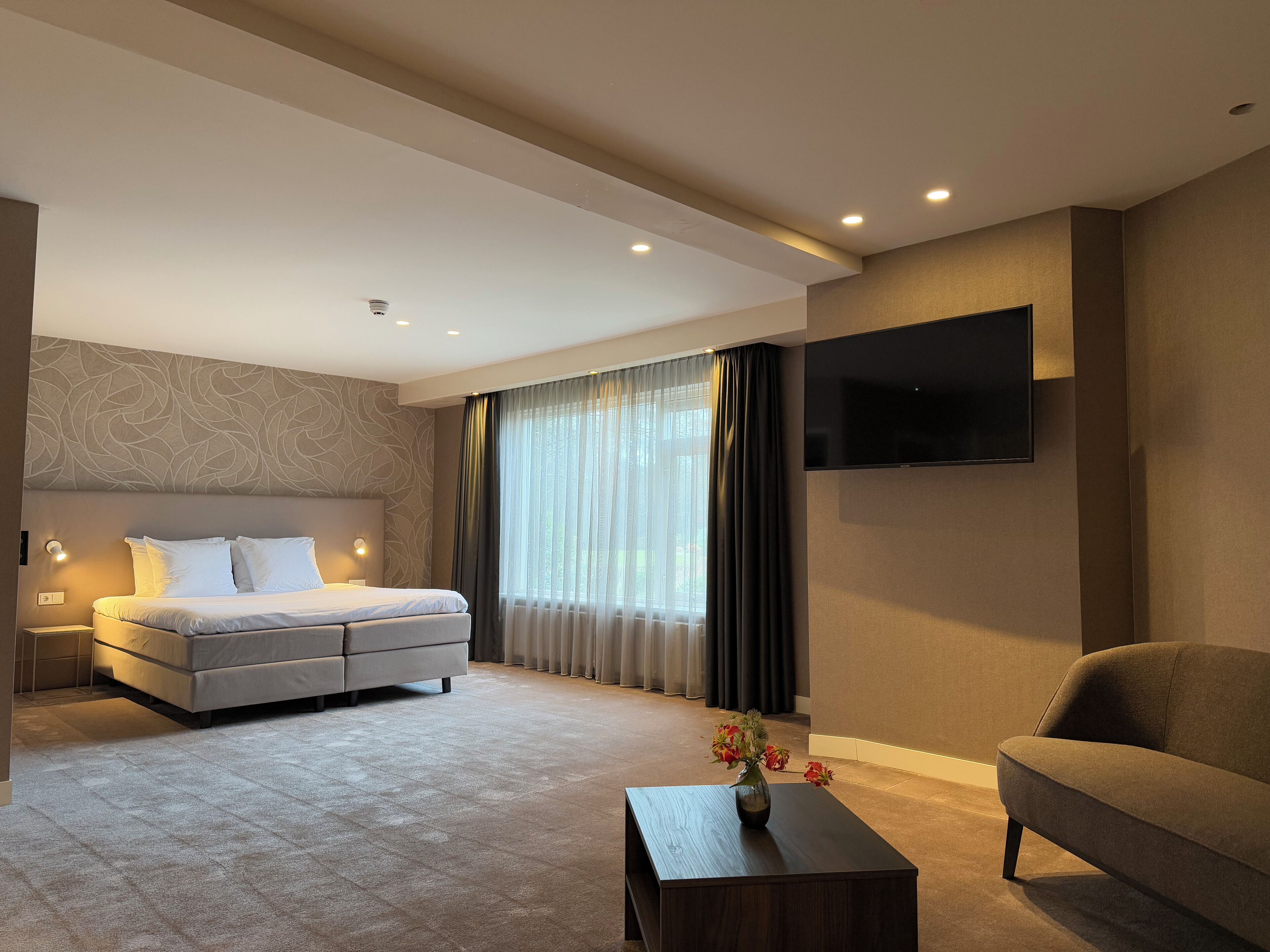 Une chambre d’hôtel moderne avec un grand lit, un canapé, une table basse agrémentée de fleurs, un téléviseur fixé au mur et un mur à motifs.