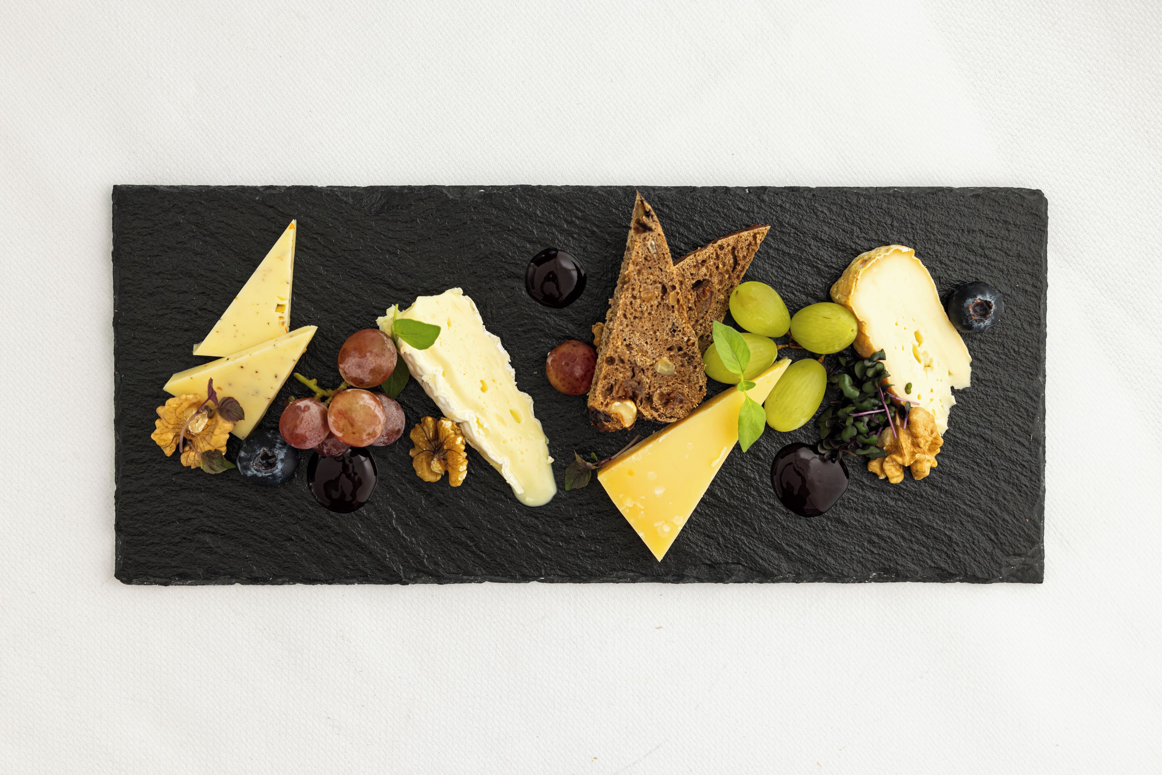 A black slate board with various cheeses, grapes, nuts, and bread slices.