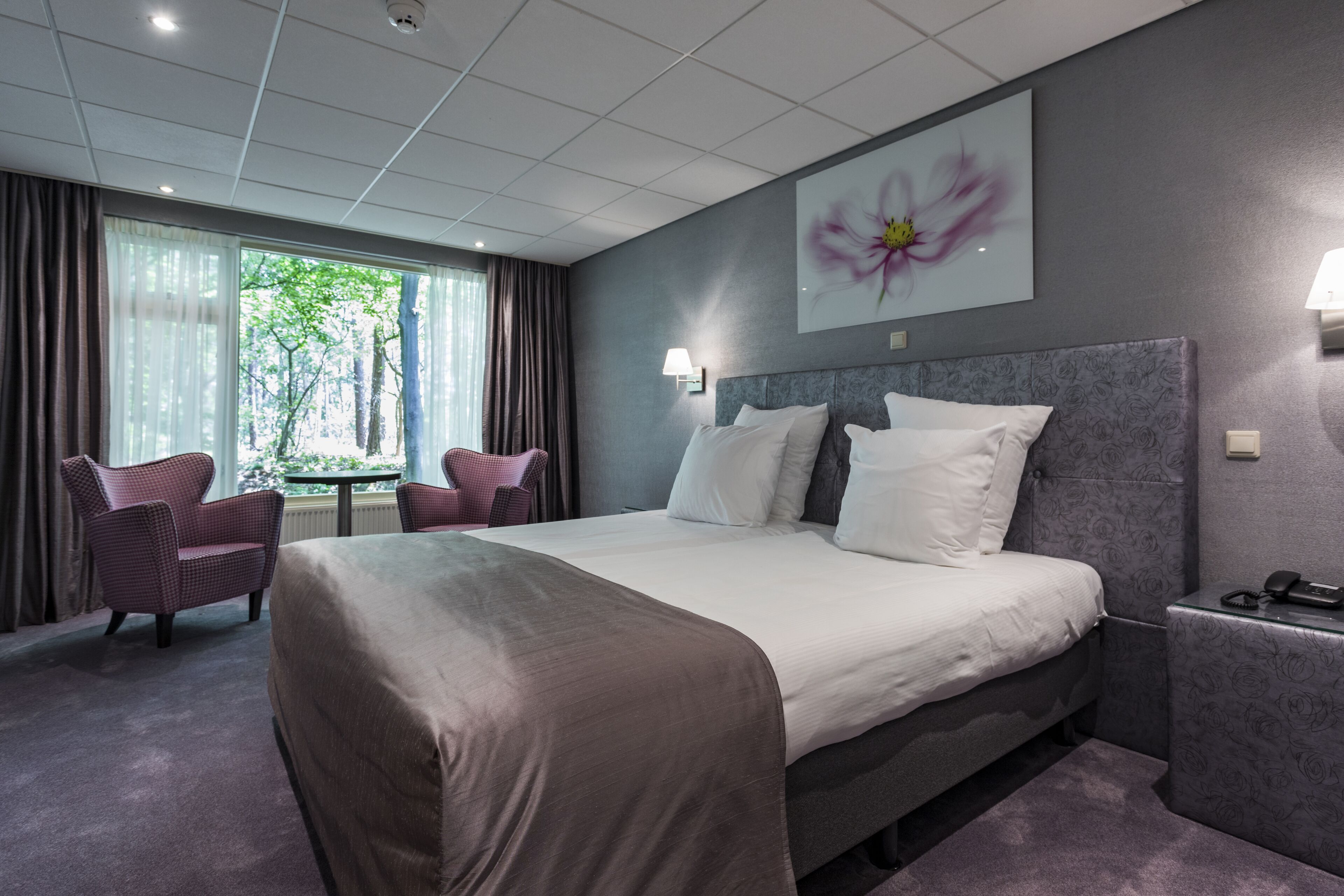 A hotel room with a large bed, two chairs, a small table, and a window with curtains.